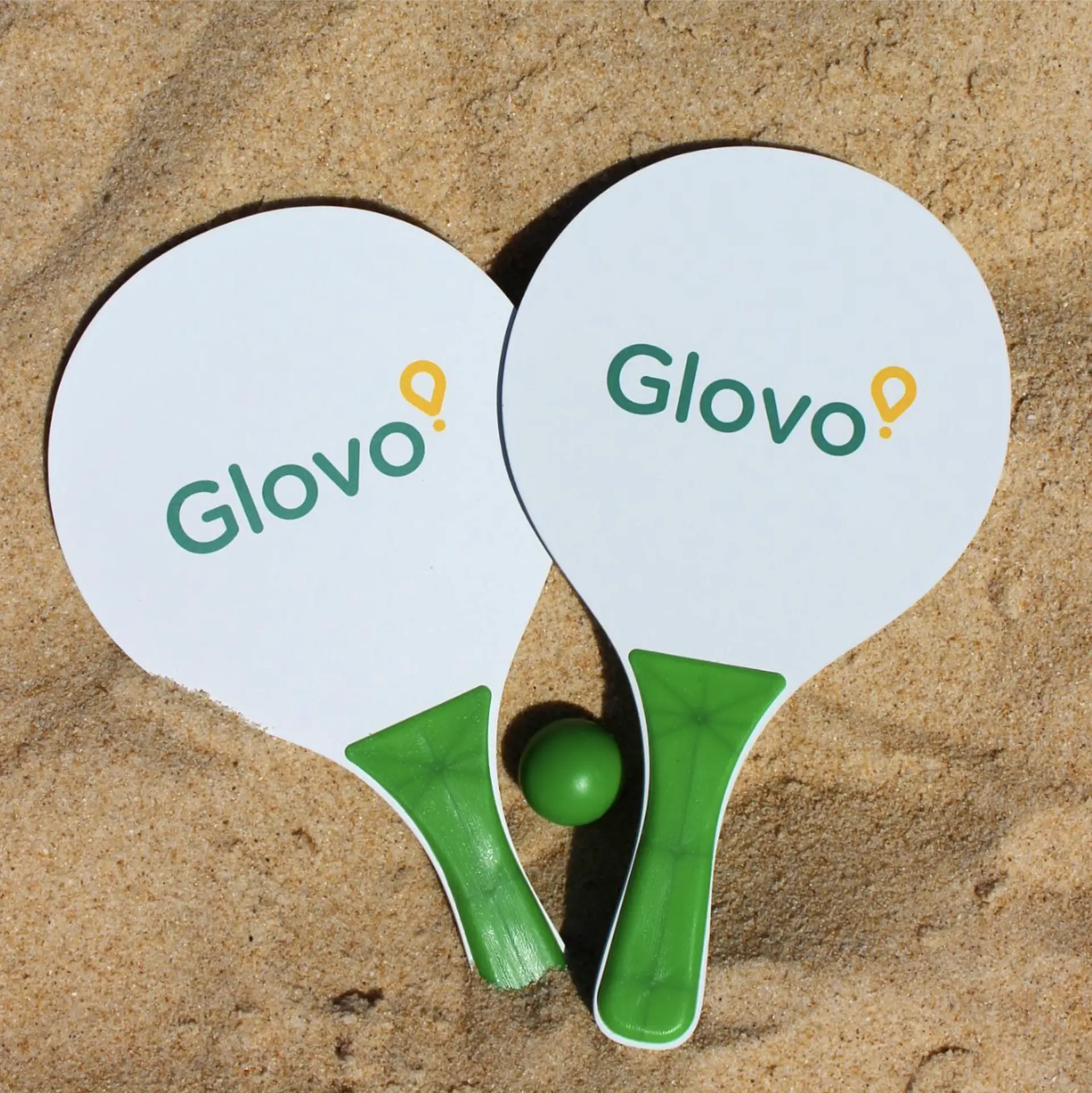 Pair of white Glovo branded beach bat rackets with green handles on sand